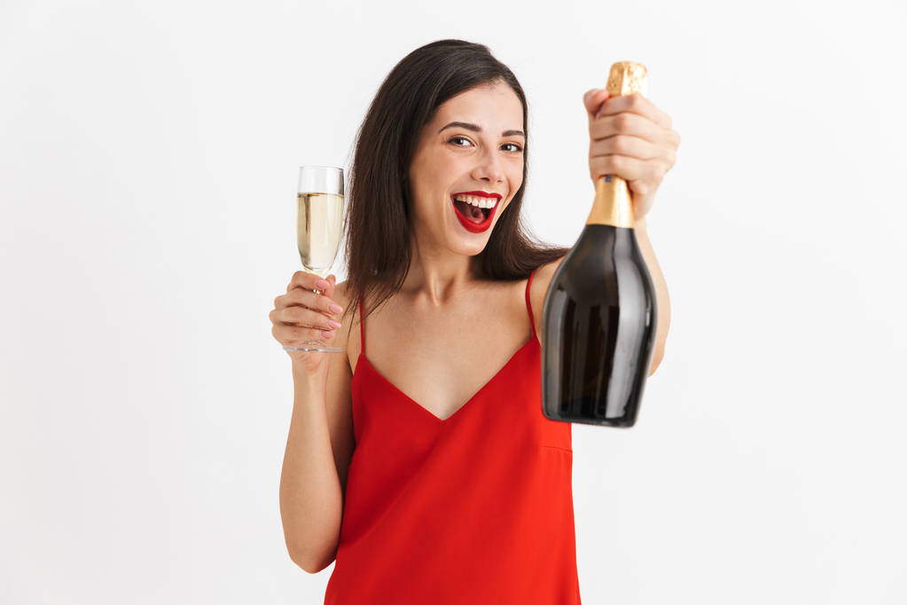 Portret van een vrolijke jonge vrouw in jurk holding glas en fles champagne geïsoleerd op witte achtergrond - Foto, afbeelding