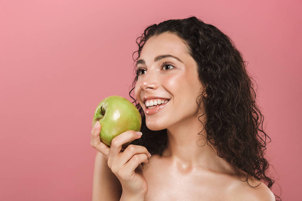 Retrato de beleza de uma jovem mulher de topless sorridente com cabelo morena encaracolado posando sobre fundo rosa, segurando maçã verde
 - Foto, Imagem