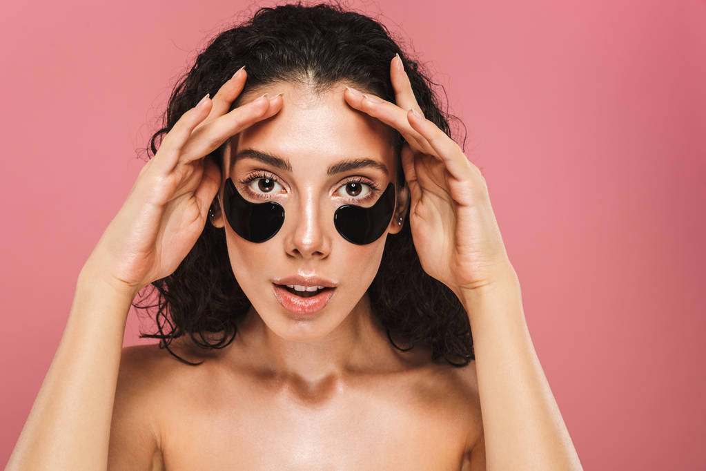 Portrait of european shirtless woman with long brunette hair wearing cosmetic patches under her eyes isolated over pink background - Photo, Image