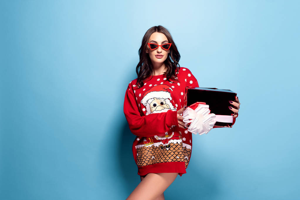 hübsche brünette Frau in Sonnenbrille und rotem Oversize-Pullover mit Weihnachtsmann-Design mit Geschenkschachtel auf blau - Foto, Bild