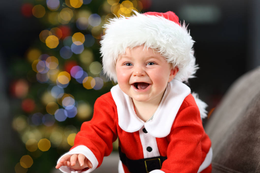 Joyful baby wears santa claus costume on a couch at home in christmas with a tree in the background - Photo, Image