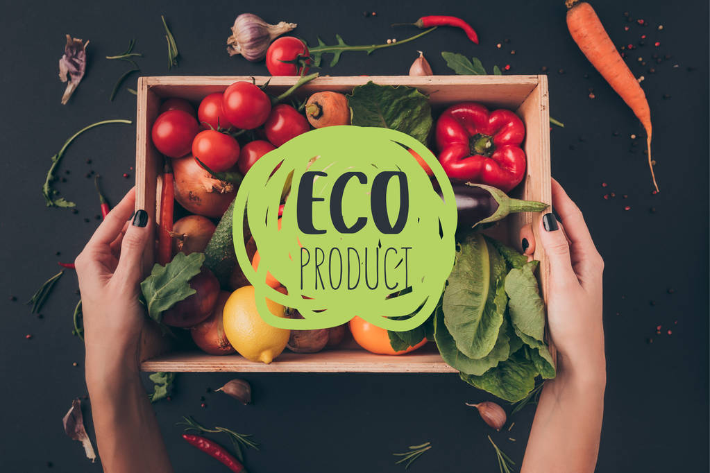 обрізане зображення жінки, що тримає дерев'яну коробку з овочами над сірим столом з написом "еко-продукт"
 - Фото, зображення