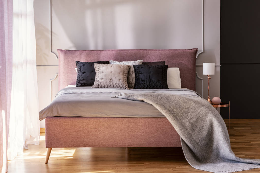 Grijze deken en zwarte kussens op roze bed in hotel slaapkamer interieur met lamp op tafel. Echte foto - Foto, afbeelding