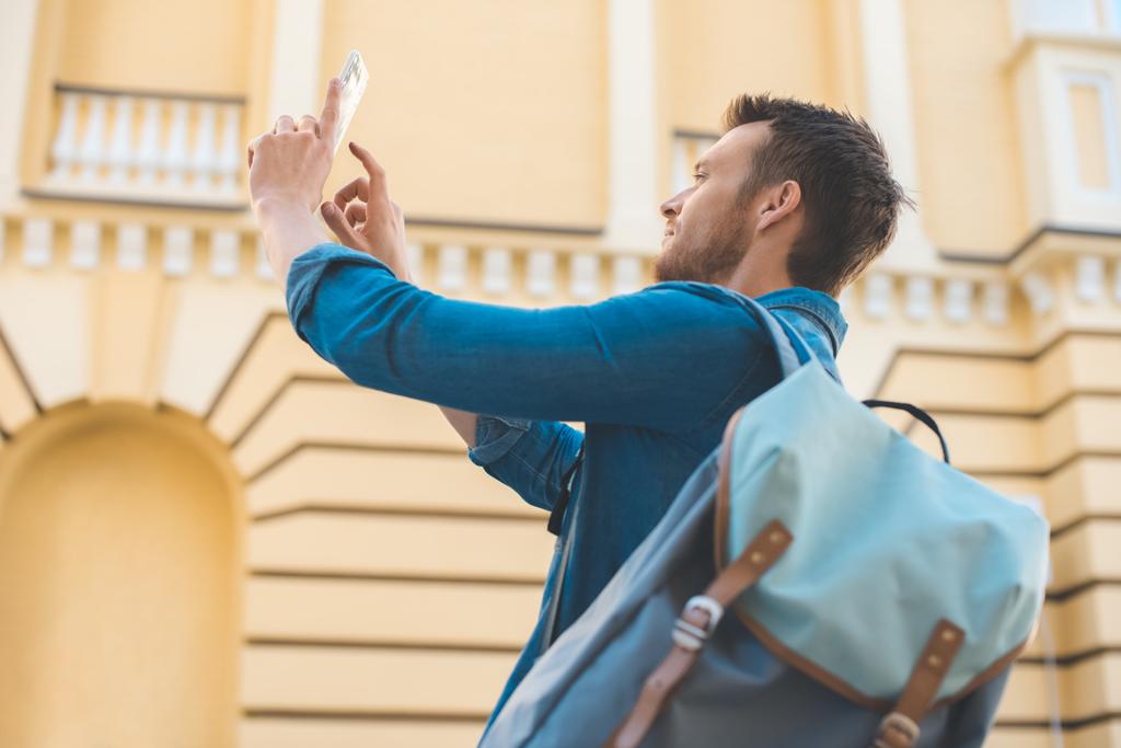 красивий молодий турист з рюкзаком фотографує зі смартфоном на вулиці
 - Фото, зображення
