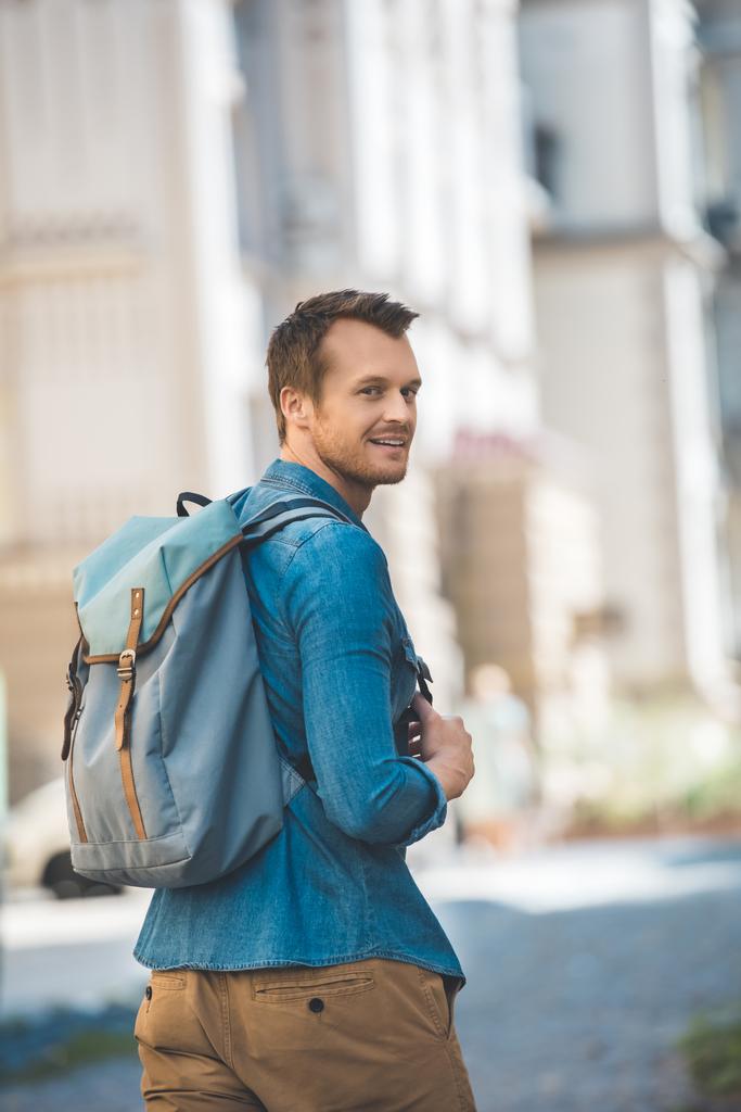 usměvavý mladý muž s batohem walking Street a při pohledu na fotoaparát - Fotografie, Obrázek