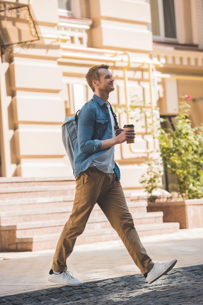casual young man with coffee to go and backpack walking by street and looking away - Photo, Image