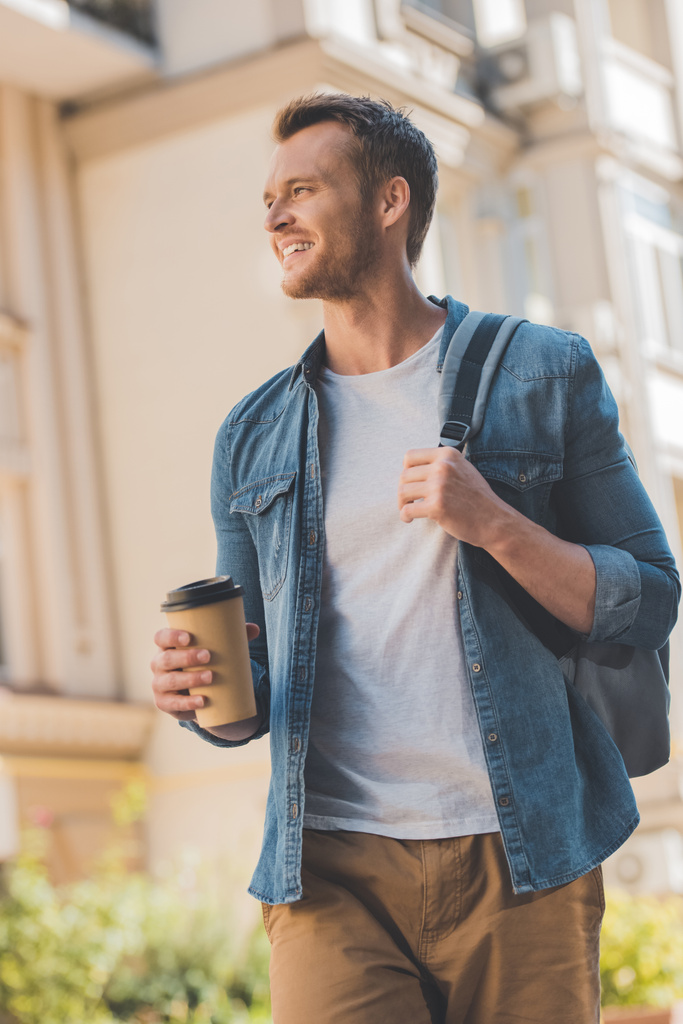 bottom view of happy young man with coffee to go and backpack walking on street and looking away - Photo, Image