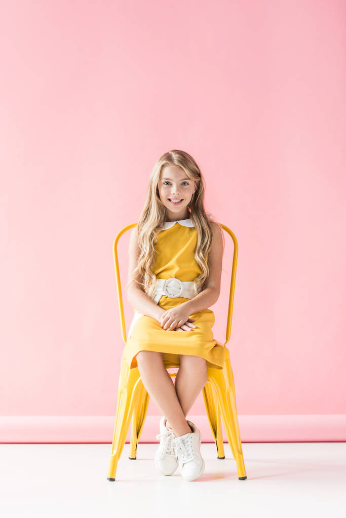 happy fashionable adorable youngster sitting on yellow chair on pink - Photo, Image