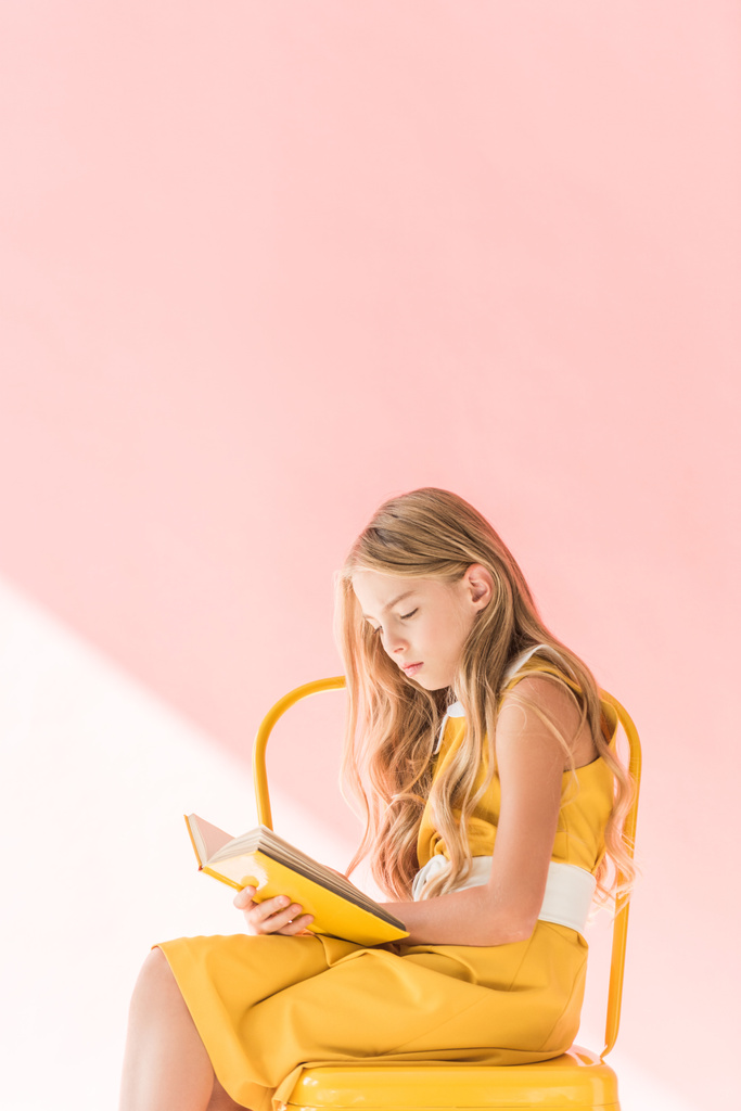 stylish blonde child reading book while sitting on yellow chair on pink - Photo, Image