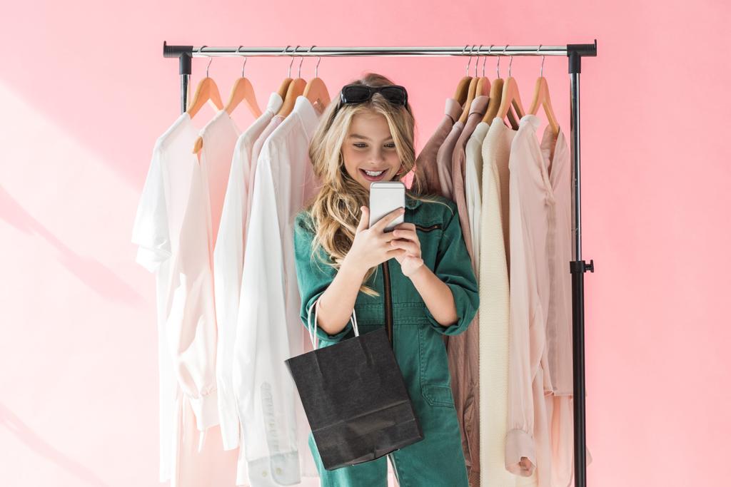 vidám gyermek divatos bevásárló táskát használ smartphone áruház közelében ruhák  - Fotó, kép