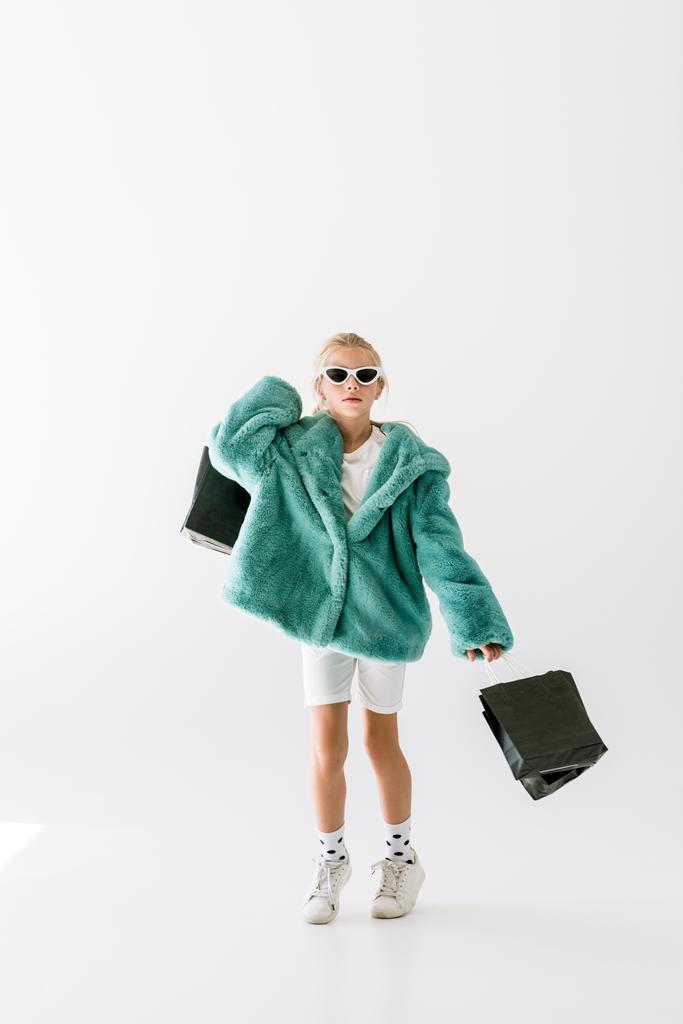 schattig stijlvolle kind in turquoise jas en zonnebril springen met zwarte boodschappentassen geïsoleerd op wit - Foto, afbeelding