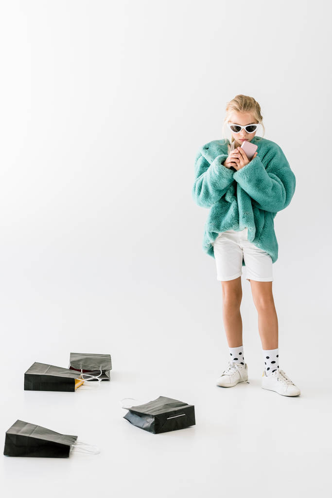 стильна дитина в бірюзовому шубці з використанням смартфона на білому з чорними сумками
 - Фото, зображення