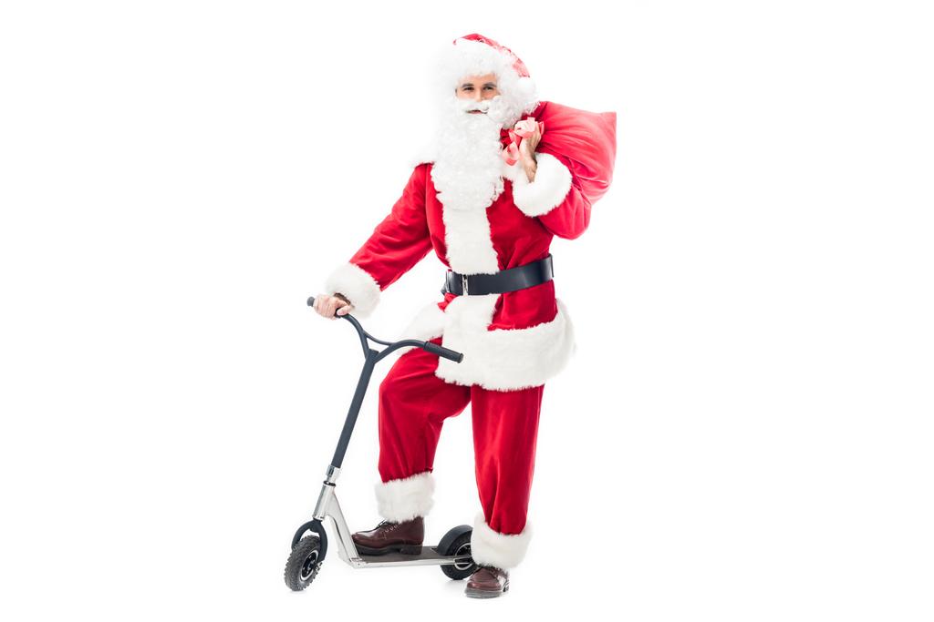Santa claus in kostuum permanent op kick scooter en chirstmas zak houden over schouder geïsoleerd op witte achtergrond - Foto, afbeelding