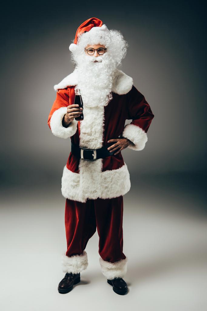 glücklich Weihnachtsmann im Kostüm stehend mit Sahne-Limo-Flasche auf grauem Hintergrund  - Foto, Bild