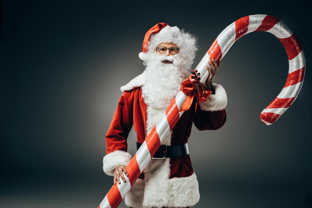 grave Santa Claus en costume debout avec gros bâton de Noël rayé isolé sur fond gris
  - Photo, image