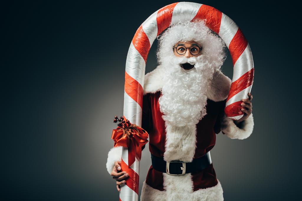 verbaasd kerstman kostuum reputatie met grote gestreepte kerst stick geïsoleerd op een grijze achtergrond  - Foto, afbeelding