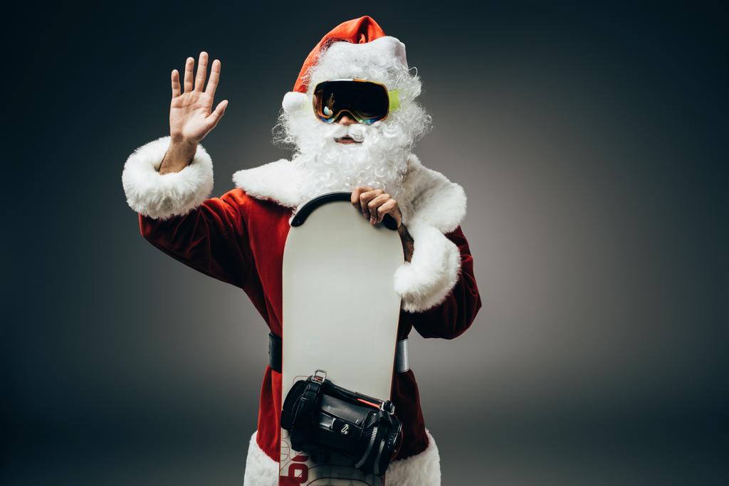 Weihnachtsmann in Skimaske steht mit Snowboard und winkt von Hand isoliert auf grauem Hintergrund  - Foto, Bild