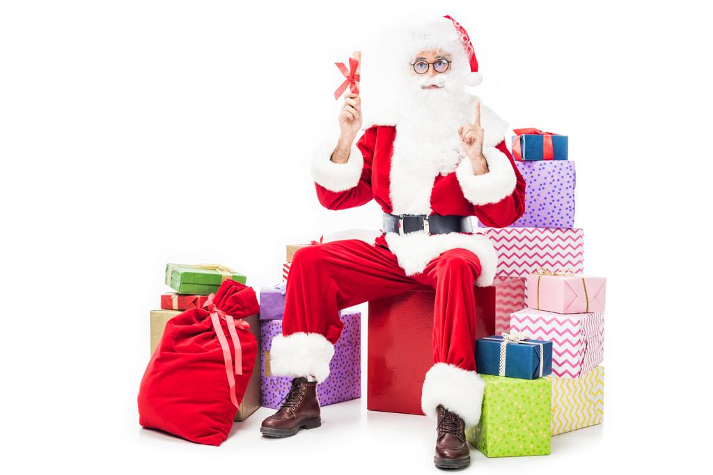 Weihnachtsmann macht Idee Geste und hält Weihnachtsgeschenk, während er auf einem Stapel von Geschenkboxen auf weißem Hintergrund isoliert sitzt  - Foto, Bild