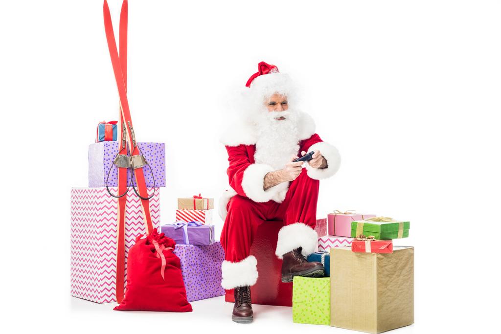 santa claus sitting on pile of gift boxes playing video game by joystick isolated on white background  - Photo, Image