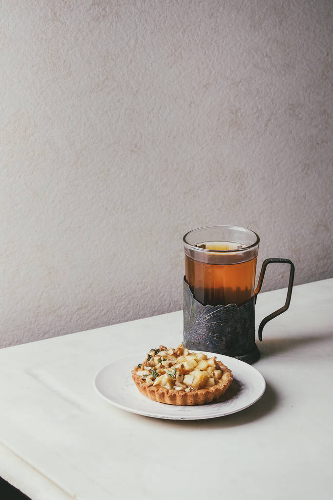Tartaleta casera de pan corto de manzana dulce en plato blanco, vaso de té caliente en portavasos vintage sobre mesa de mármol blanco. Horneado de otoño. Estilo minimalista
. - Foto, Imagen