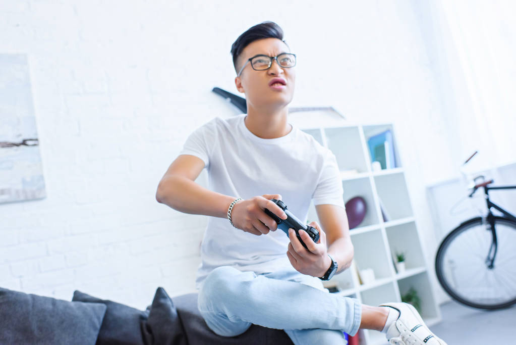 роздратований азіатський чоловік грає у відеогру з джойстиком на дивані вдома
 - Фото, зображення