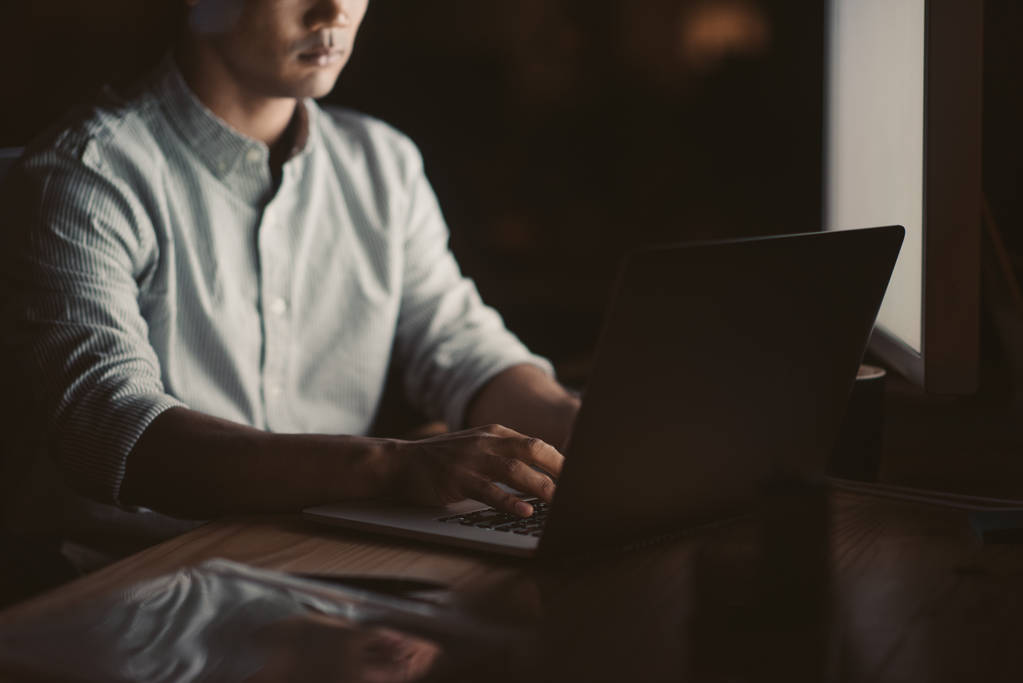 Primer plano de un hombre de negocios que trabaja en línea con un ordenador portátil mientras está sentado en su escritorio en una oficina oscura tarde en la noche
 - Foto, imagen