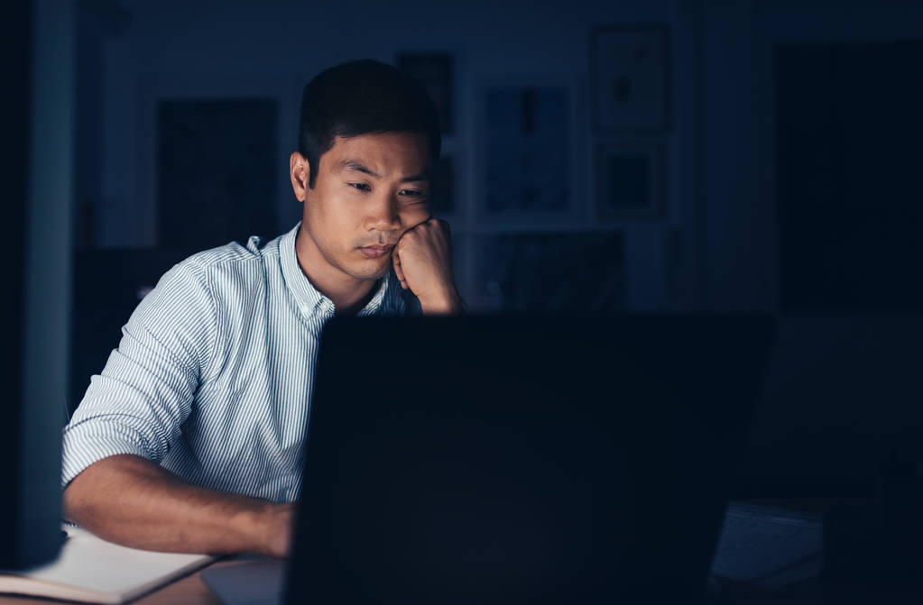 müde junge asiatische Geschäftsmann sitzt an seinem Schreibtisch und arbeitet an einem Laptop in einem dunklen Büro spät am Abend - Foto, Bild