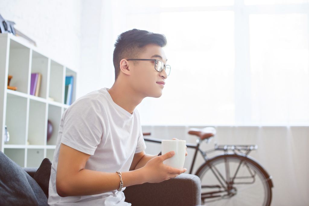 Seitenansicht eines hübschen asiatischen Mannes, der morgens zu Hause auf dem Sofa sitzt und eine Tasse Tee hält - Foto, Bild