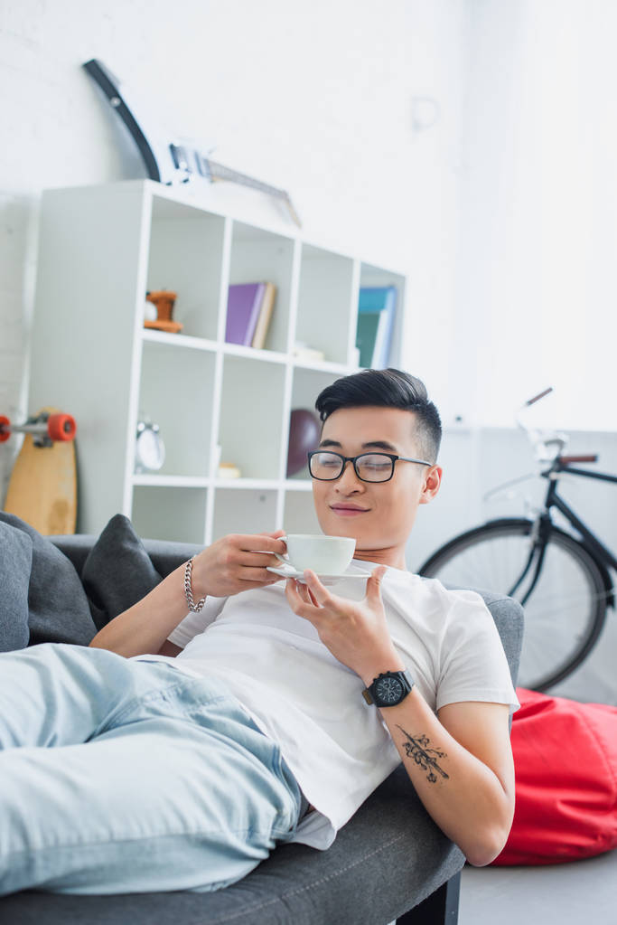 szczęśliwy młody człowiek Azjatka w okularach, leżąc na kanapie i trzymając kubek kawy - Zdjęcie, obraz