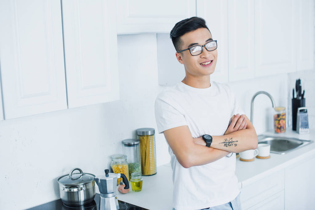 pohledný mladý usmívající se asijské muž v brýlích s překřížením rukou v kuchyni - Fotografie, Obrázek