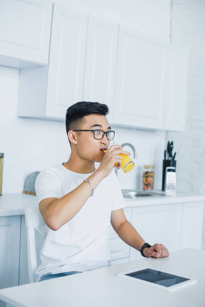 junger asiatischer Mann in Brille trinkt Saft und schaut in Küche weg - Foto, Bild