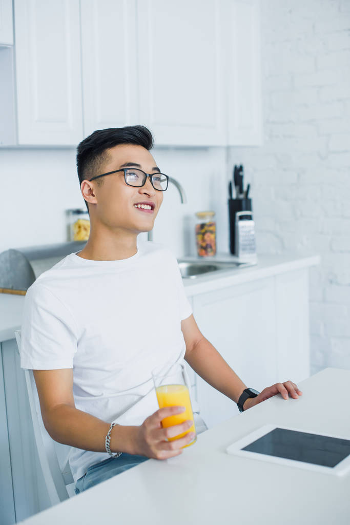komea hymyilevä nuori aasialainen mies silmälasit tilalla lasi fo mehua ja katselee pois, kun istuu keittiössä
 - Valokuva, kuva