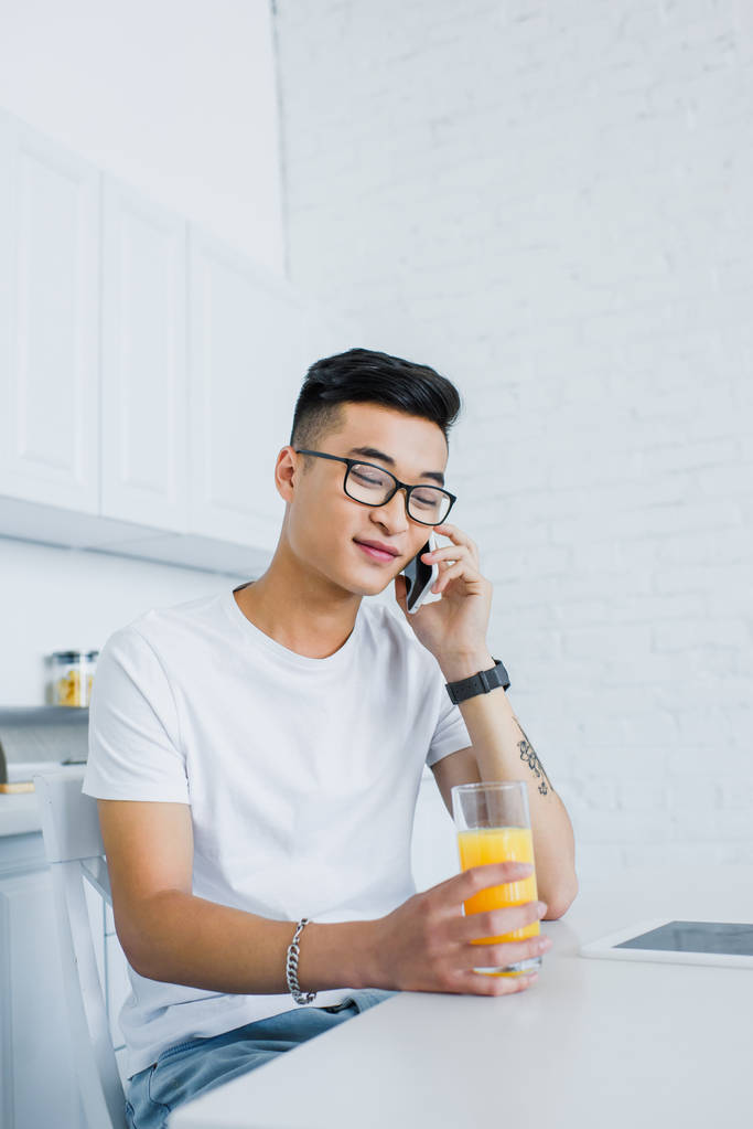 młody człowiek Azjatka w okularach, trzymając szklankę soku i rozmawiać przez smartfona, siedząc w kuchni - Zdjęcie, obraz