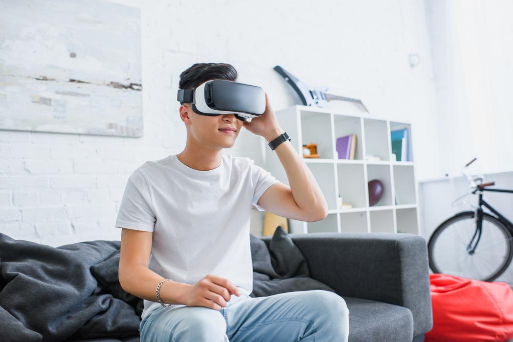 mosolygó fiatal ázsiai ember segítségével virtuális-valóság sisak, miközben otthon a fotelben ülve - Fotó, kép