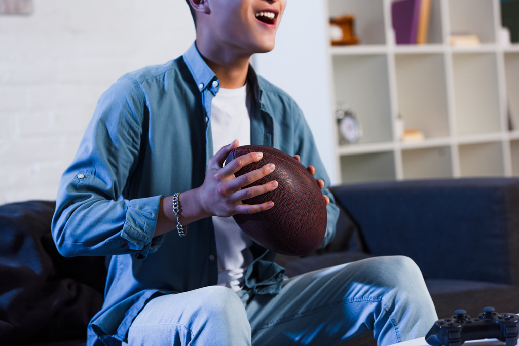 Schnappschuss eines lächelnden jungen Mannes, der Rugbyball in der Hand hält und sich zu Hause ein Sportspiel anschaut - Foto, Bild