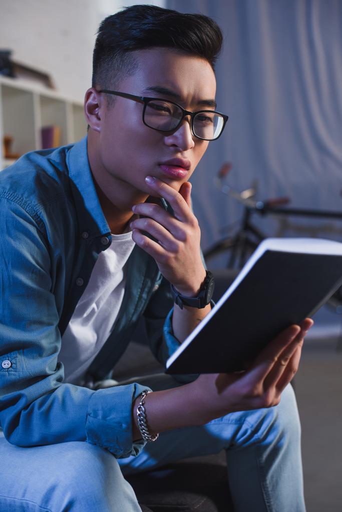 focalizzato giovane asiatico uomo in occhiali lettura libro a casa
 - Foto, immagini