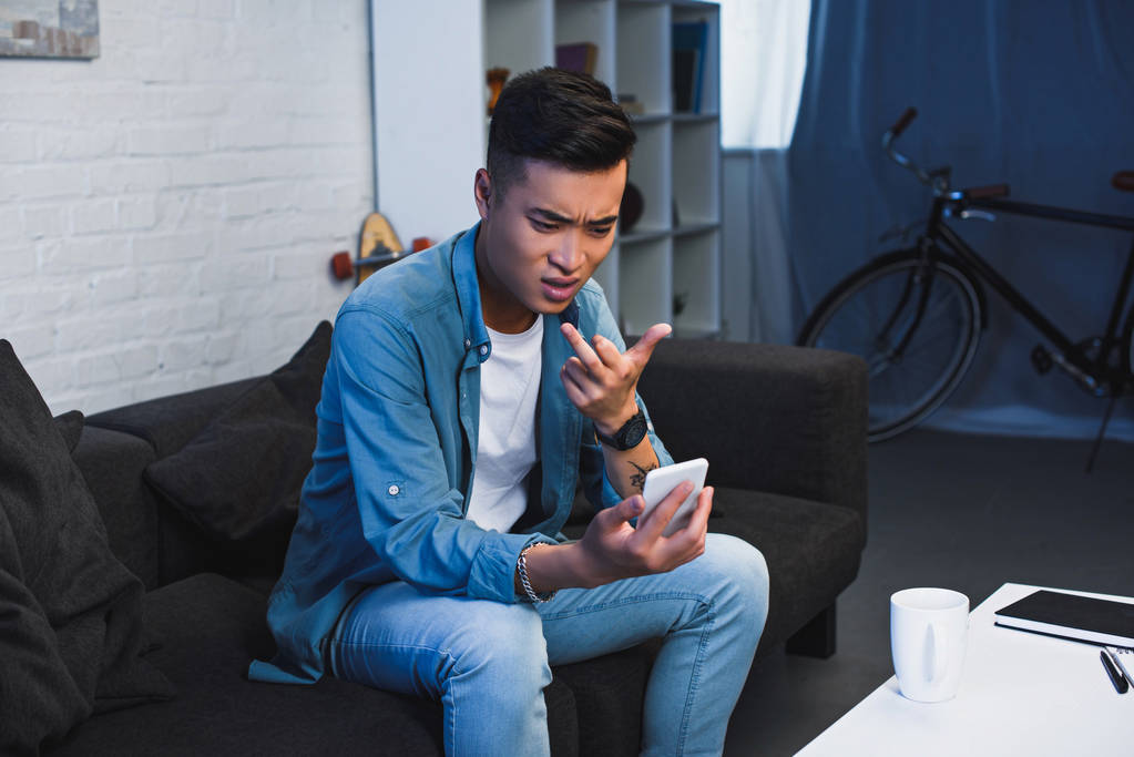 молодой азиат, использующий смартфон и показывающий средний палец
 - Фото, изображение