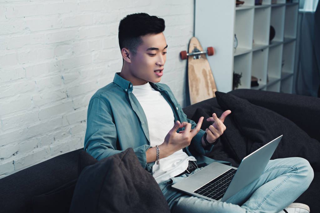 junge asiatische Mann sitzt auf Couch und gibt Mittelfinger auf Laptop - Foto, Bild