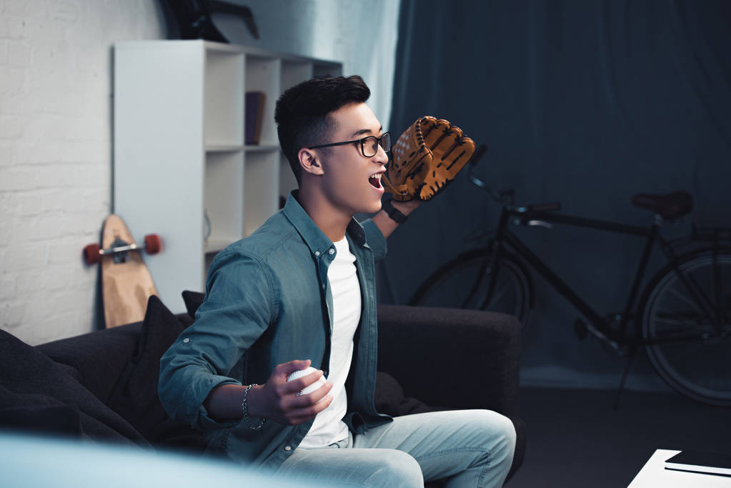 emocjonalne młode Azji człowiek z rękawicy baseballowej i piłka siedzi na kanapie i oglądać sport mecz w domu   - Zdjęcie, obraz