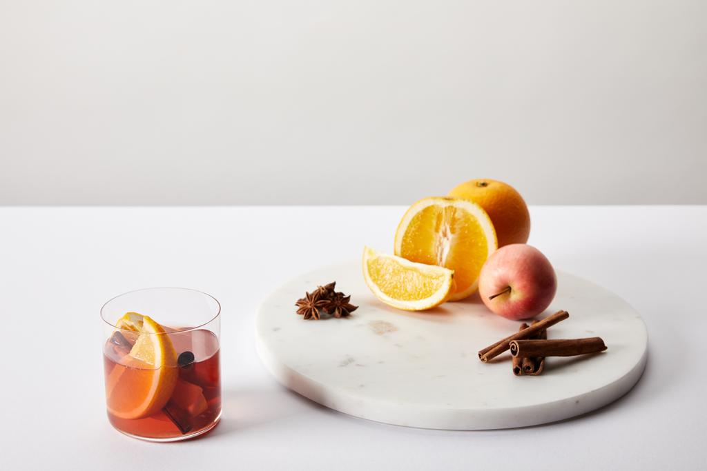 Nahaufnahme von Glühwein im Glas, Gewürzen, Orangen und Äpfeln auf weißer Oberfläche vor grauem Hintergrund - Foto, Bild