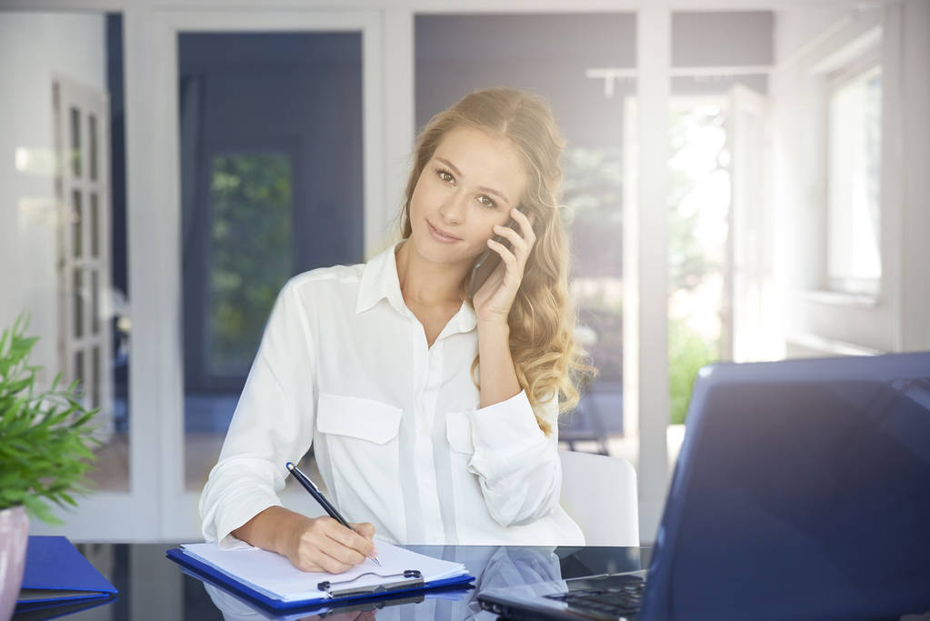 Πορτρέτο του όμορφη νεαρή Βοηθός επιχειρηματίας κάνοντας κλήση ενώ κάθεται στο γραφείο και καθιστώντας σημειώσεις. - Φωτογραφία, εικόνα