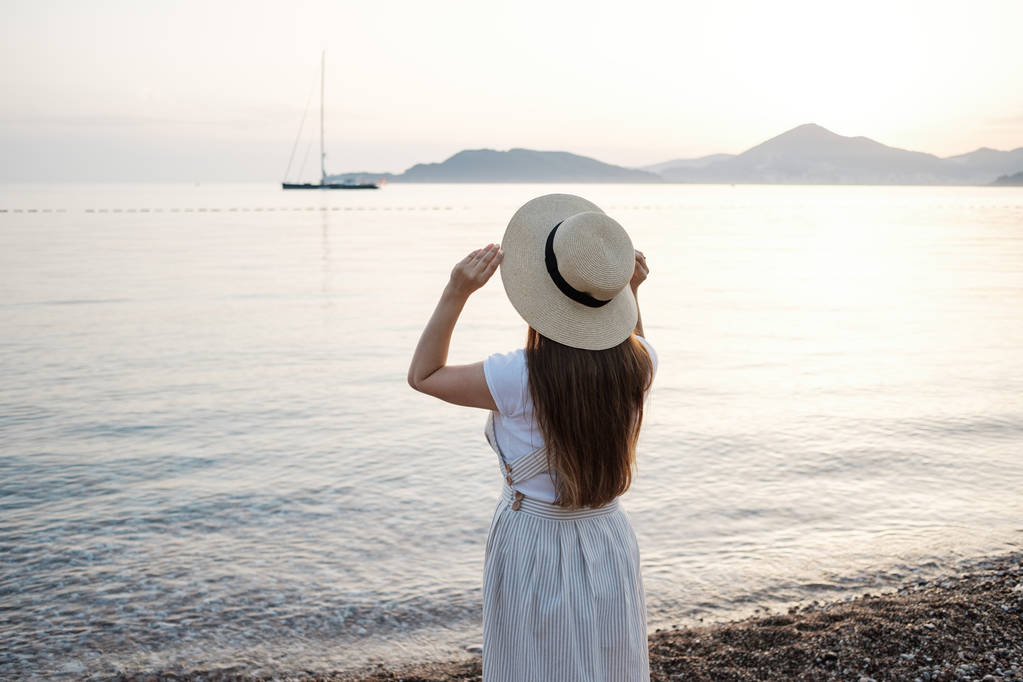 Vista trasera de una joven hermosa mujer sosteniendo su sombrero de paja en la playa y mirando al horizonte
. - Foto, Imagen