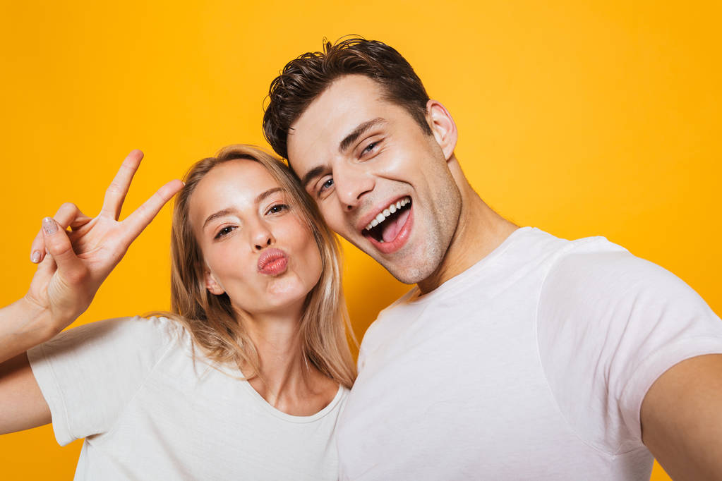 Foto de feliz animado jovem casal amoroso de pé isolado sobre fundo de parede amarela tirar uma selfie por câmera mostrando gesto de paz
. - Foto, Imagem