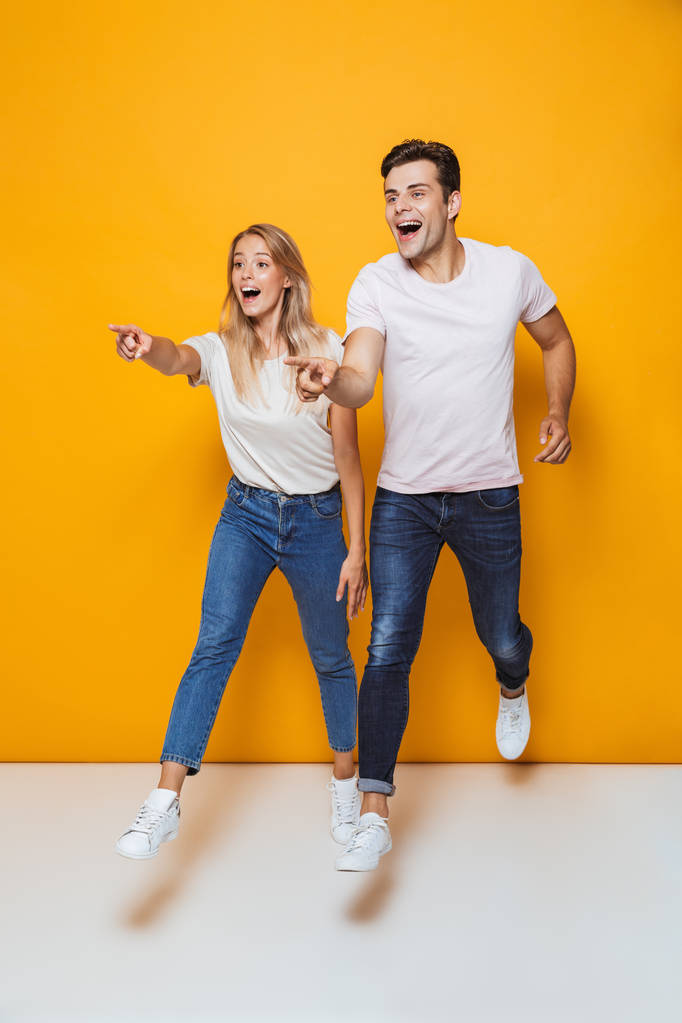 Photo of happy excited young loving couple jumping isolated over yellow wall background pointing. - Photo, Image