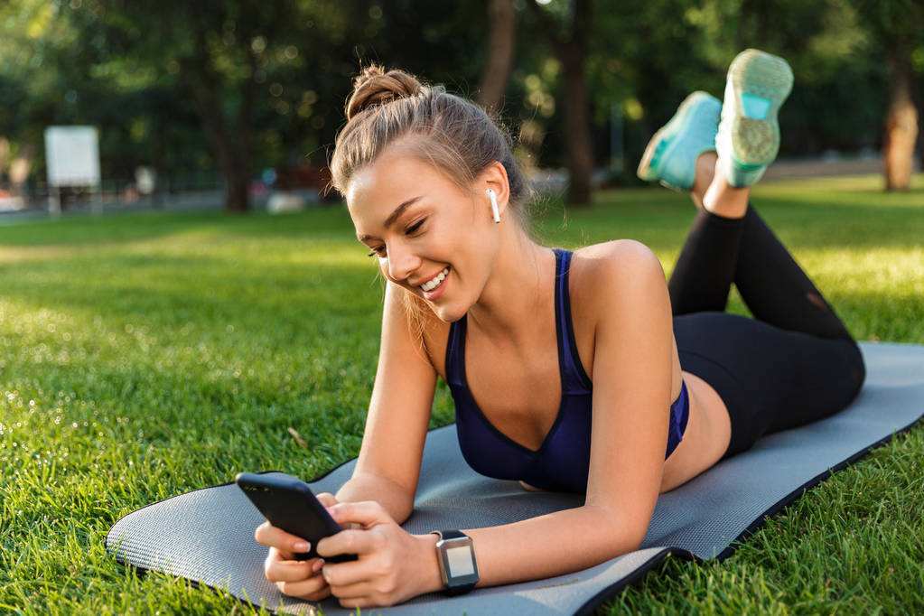 Portrait of a happy young fitness girl laying on a mat at the park, using mobile phone, listening to music with earphones - Photo, Image