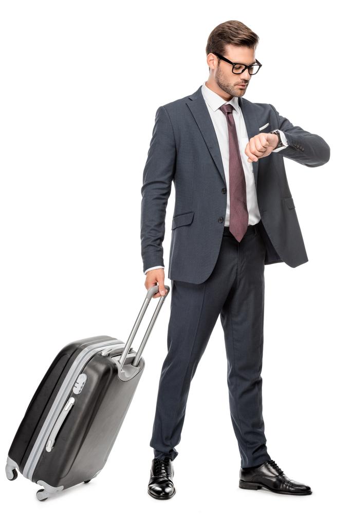 joven hombre de negocios guapo con equipaje mirando reloj de pulsera aislado en blanco
 - Foto, imagen
