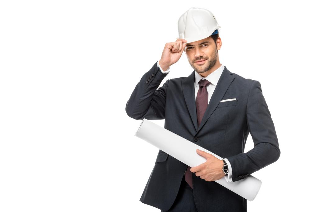 щасливий молодий чоловік архітектор в костюмі тримає креслення і регулює жорсткий капелюх ізольовано на білому
  - Фото, зображення