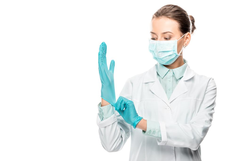 νεαρή γυναίκα γιατρό σε ιατρική μάσκα βάζοντας σε γάντια λατέξ που απομονώνονται σε λευκό - Φωτογραφία, εικόνα