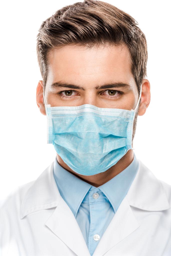 serious young male doctor in medical mask looking at camera isolated on white  - Photo, Image