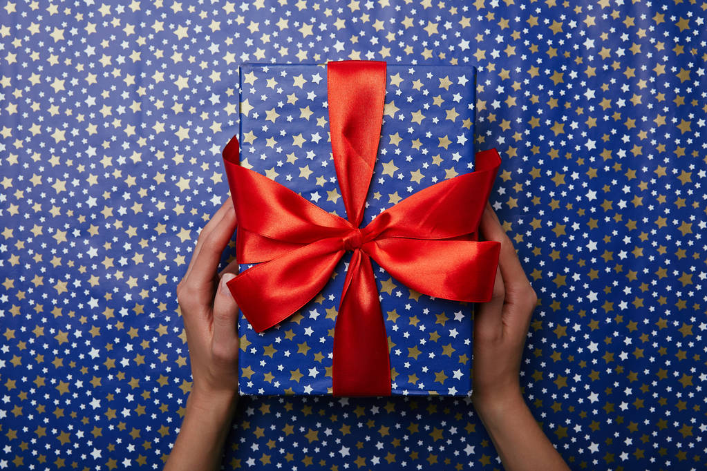 cropped shot of woman holding bright wrapped gift with red ribbon on wrapping paper with stars pattern - Photo, Image